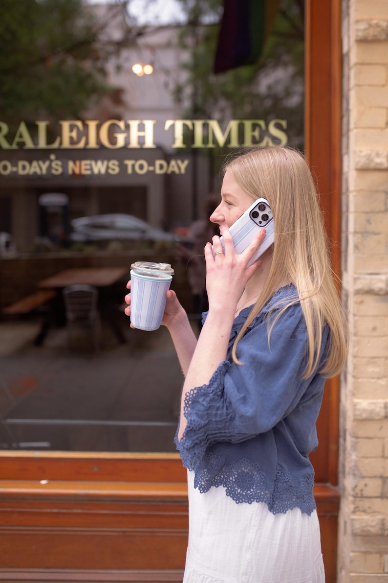 May include: A light blue and white striped reusable coffee cup with a clear lid, held alongside a white smartphone with a matching light blue and white striped case, featuring a blue blouse with scalloped lace trim and a white skirt.
