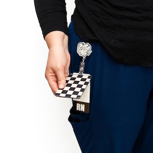 Puede incluir: Un porta-credenciales a cuadros blancos y negros con una correa de pl&aacute;stico transparente. El porta-credenciales lo sostiene una persona que lleva una camisa negra de manga larga y un uniforme azul. Un carrete de insignia RN plateado est&aacute; sujeto al uniforme.