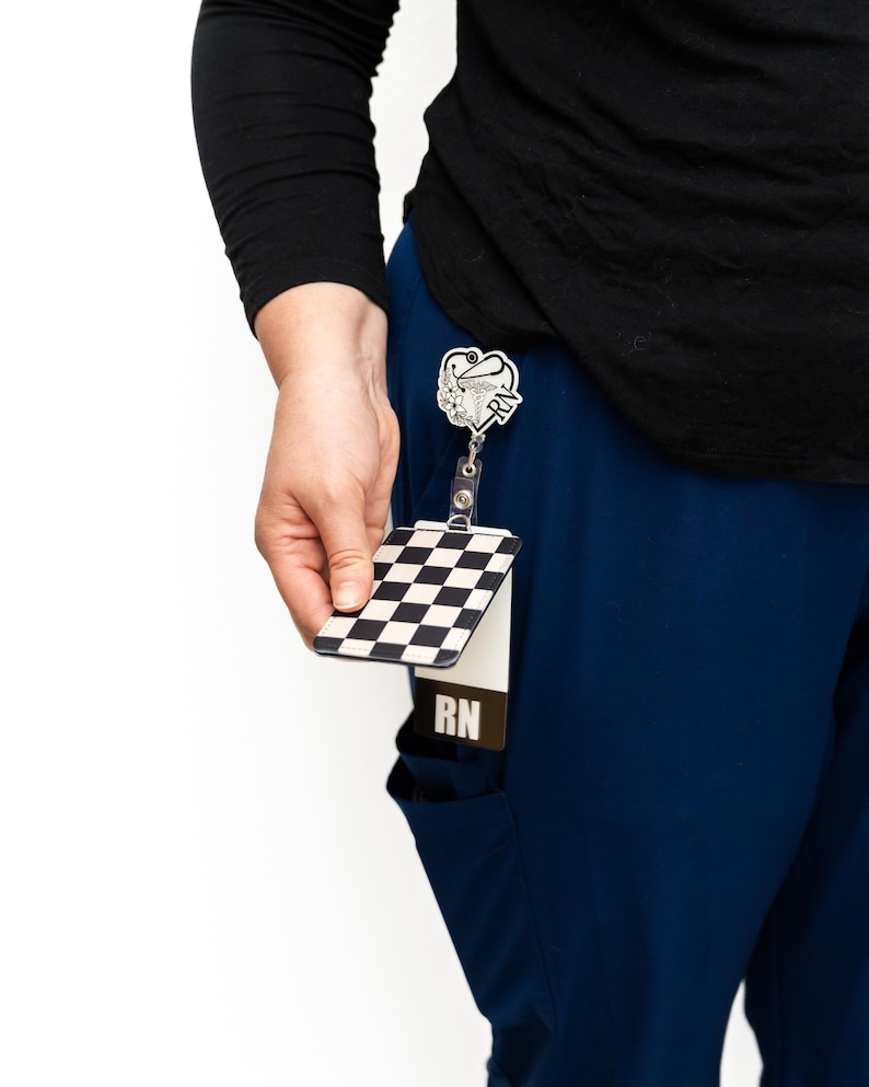 Puede incluir: Un porta-credenciales a cuadros blancos y negros con una correa de pl&aacute;stico transparente. El porta-credenciales lo sostiene una persona que lleva una camisa negra de manga larga y un uniforme azul. Un carrete de insignia RN plateado est&aacute; sujeto al uniforme.