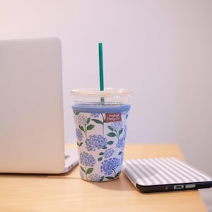 May include: A clear plastic tumbler with a teal straw and a light blue fabric sleeve featuring a blue and white hydrangea floral pattern. A small brown rectangular tag with the text Frederick Family Co is attached to the sleeve.