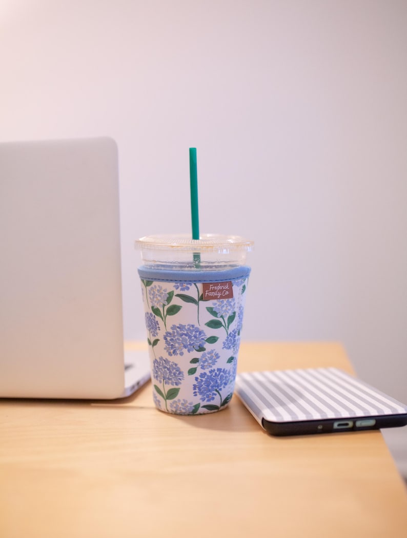 May include: A clear plastic tumbler with a teal straw and a light blue fabric sleeve featuring a blue and white hydrangea floral pattern. A small brown rectangular tag with the text Frederick Family Co is attached to the sleeve.