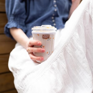 May include: A cylindrical beverage cup sleeve featuring a light beige and white vertical striped pattern with small orange floral accents. A small rectangular brown fabric tag with the text Frederick Family Co is stitched onto the upper front.