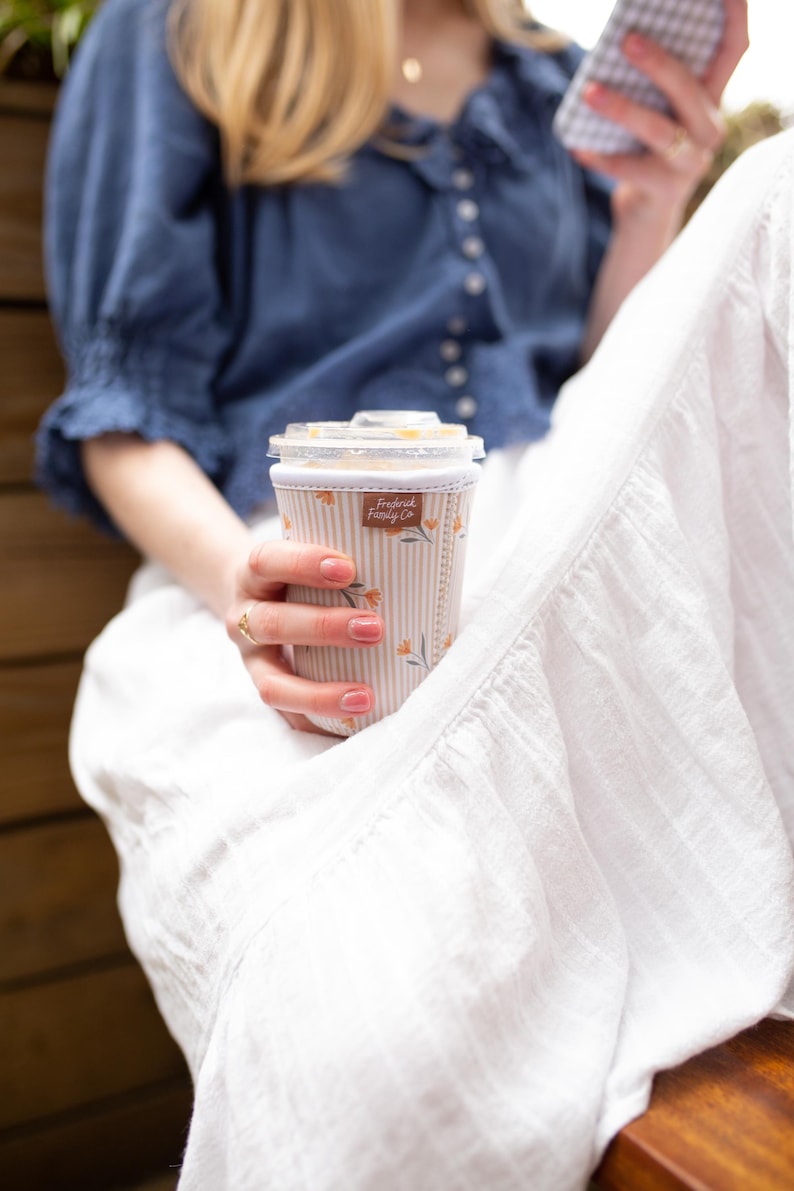 May include: A cylindrical beverage cup sleeve featuring a light beige and white vertical striped pattern with small orange floral accents. A small rectangular brown fabric tag with the text Frederick Family Co is stitched onto the upper front.