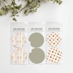 May include: Three sets of two circular car coasters in clear plastic packaging. The first set features a vertical floral pattern, the second a green and white gingham check, and the third a pattern of small pumpkins. Each label reads CAR COASTERS.