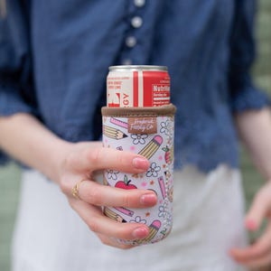 May include: A white and pink insulated can cooler with a school-themed design, featuring pencils, apples, and flowers. The cooler holds a red and white can. The brand name "Frederick Family Co." is visible on the cooler.