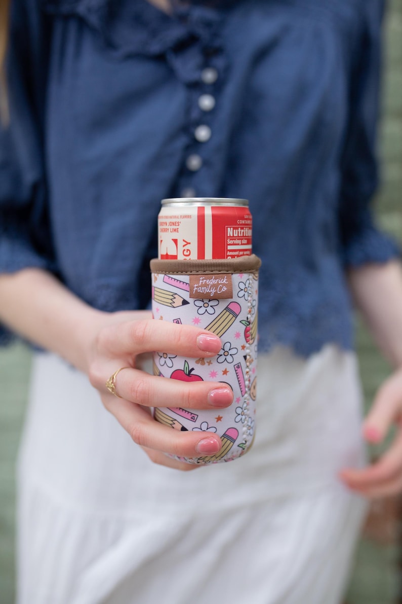 May include: A white and pink insulated can cooler with a school-themed design, featuring pencils, apples, and flowers. The cooler holds a red and white can. The brand name "Frederick Family Co." is visible on the cooler.