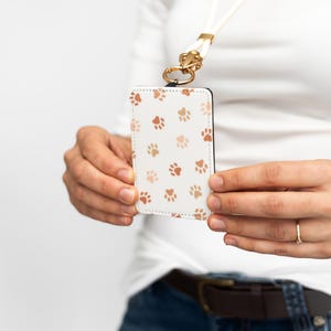Puede incluir: Portatarjetas de identificaci&oacute;n blanco con un patr&oacute;n de huellas de patas marrones. El soporte est&aacute; sujeto a un cord&oacute;n blanco con un cierre dorado. El soporte lo sostiene una persona que lleva una camisa blanca y vaqueros azules.
