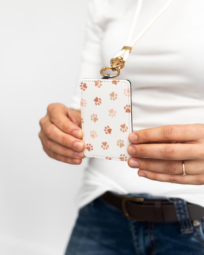 Puede incluir: Portatarjetas de identificaci&oacute;n blanco con un patr&oacute;n de huellas de patas marrones. El soporte est&aacute; sujeto a un cord&oacute;n blanco con un cierre dorado. El soporte lo sostiene una persona que lleva una camisa blanca y vaqueros azules.