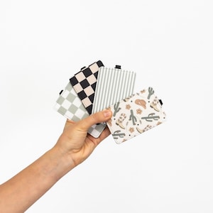 Puede incluir: Una mano sostiene una colecci&oacute;n de tarjeteros estampados. Los dise&ntilde;os incluyen cuadros blancos y negros, rayas grises y blancas y un estampado crema con botas de vaquero, cactus y sombreros. Cada tarjetero tiene una peque&ntilde;a pesta&ntilde;a negra.