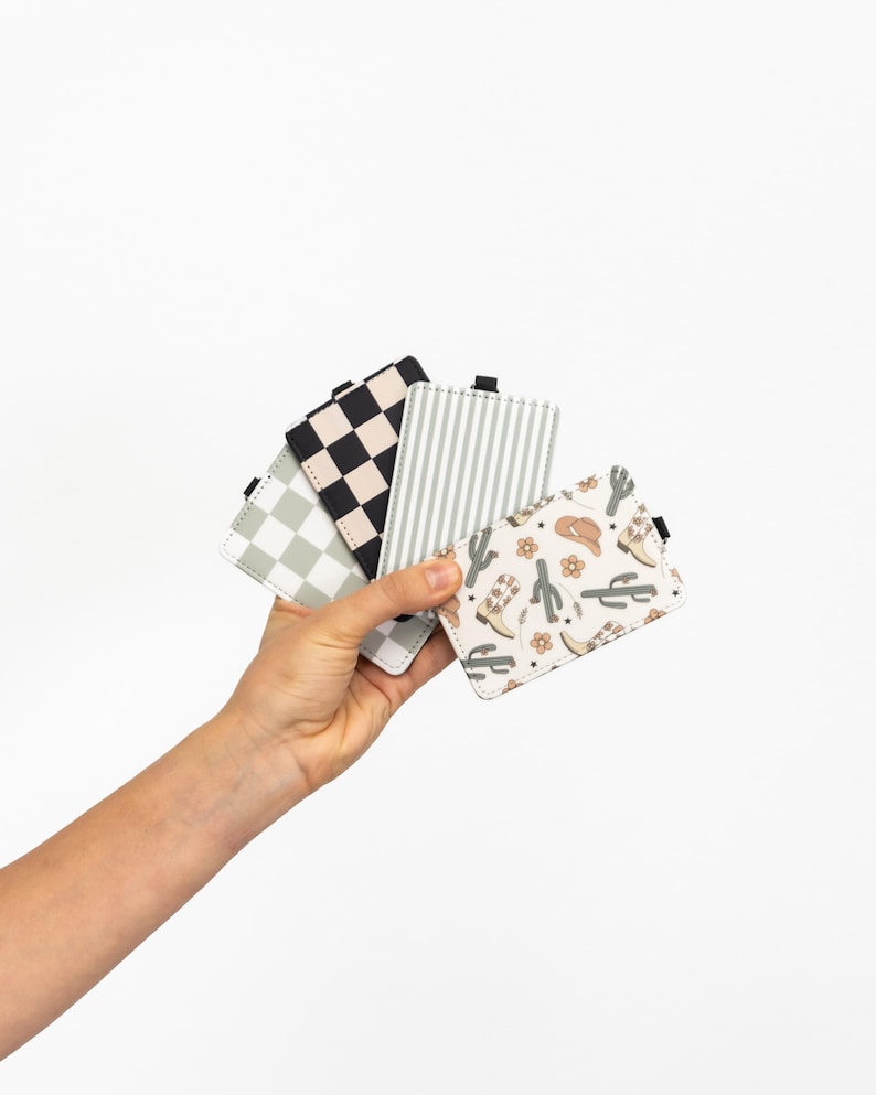 Puede incluir: Una mano sostiene una colecci&oacute;n de tarjeteros estampados. Los dise&ntilde;os incluyen cuadros blancos y negros, rayas grises y blancas y un estampado crema con botas de vaquero, cactus y sombreros. Cada tarjetero tiene una peque&ntilde;a pesta&ntilde;a negra.