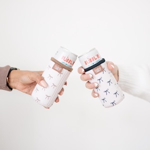 May include: Two white beverage can coolers with a bow pattern are held up. The coolers have a brown and black band around the top. The cans inside say "Bubbly" in red and blue text. The coolers are designed to keep drinks cold.