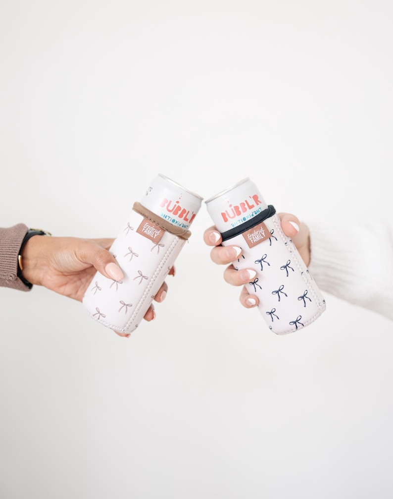 May include: Two white beverage can coolers with a bow pattern are held up. The coolers have a brown and black band around the top. The cans inside say "Bubbly" in red and blue text. The coolers are designed to keep drinks cold.