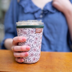 May include: A cylindrical beverage cup sleeve featuring a light pink background with a repeating pattern of small pink flowers and dark green stems. The sleeve has a dark green top trim and a small brown fabric tag labeled Frederick Family Co.