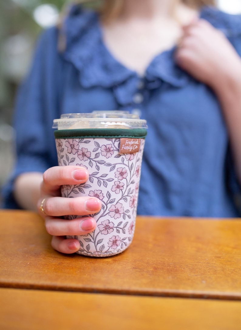 May include: A cylindrical beverage cup sleeve featuring a light pink background with a repeating pattern of small pink flowers and dark green stems. The sleeve has a dark green top trim and a small brown fabric tag labeled Frederick Family Co.