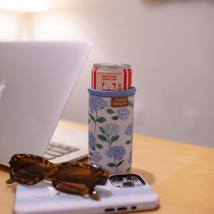 May include: A white and blue floral print can cooler with a red and white can inside. The can cooler has a blue rim and the text "Frederick Family Co." The image also includes sunglasses and a phone.