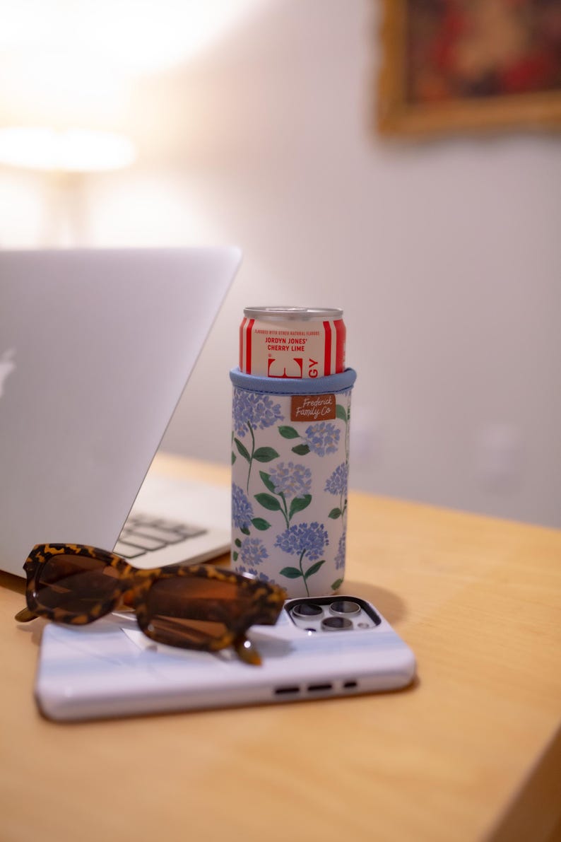 May include: A white and blue floral print can cooler with a red and white can inside. The can cooler has a blue rim and the text "Frederick Family Co." The image also includes sunglasses and a phone.