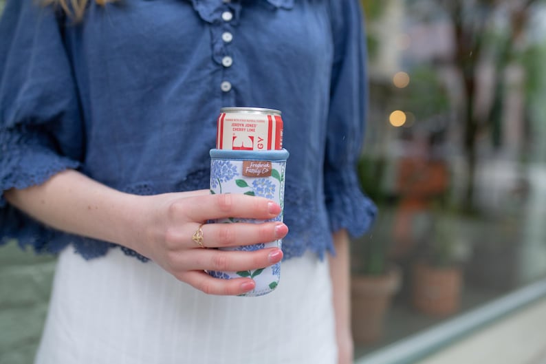 May include: A white and blue floral print can cooler holding a red and white soda can. The can cooler has a blue trim and a small label that reads "Fresh Fields Co."