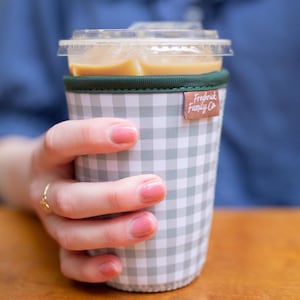 May include: A reusable cup sleeve featuring a grey and white gingham check pattern with a dark green fabric trim at the top edge. A small brown rectangular tag with the white text Frederick Family Co is attached to the side of the sleeve.