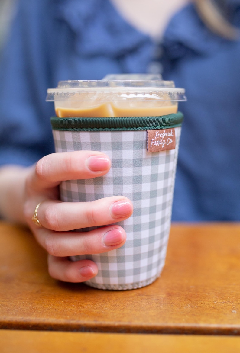 May include: A reusable cup sleeve featuring a grey and white gingham check pattern with a dark green fabric trim at the top edge. A small brown rectangular tag with the white text Frederick Family Co is attached to the side of the sleeve.