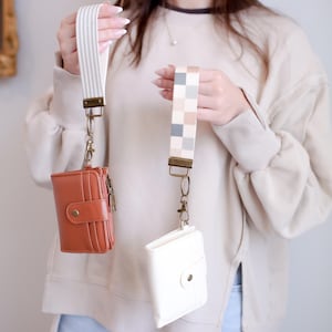 May include: Two small wallets with wrist straps are displayed. One is a brown leather wallet with a snap closure and a gold zipper. The other is a white wallet with a snap closure. The straps have different patterns.