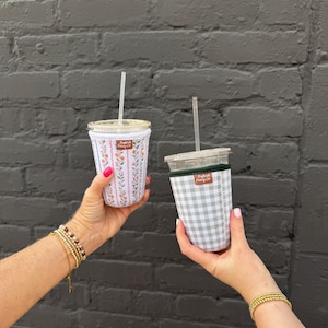 May include: Two iced coffee cups with fabric sleeves. One sleeve features a white background with vertical pink stripes and small floral patterns. The other sleeve has a light green and white gingham check pattern. Both have clear lids and straws.