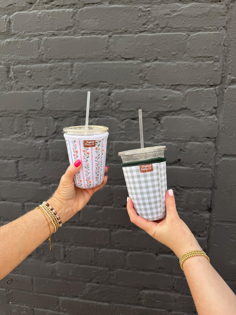 May include: Two iced coffee cups with fabric sleeves. One sleeve features a white background with vertical pink stripes and small floral patterns. The other sleeve has a light green and white gingham check pattern. Both have clear lids and straws.