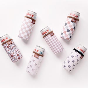 May include: Six beverage can coolers in various patterns, including checkered, floral, and bow designs. Each cooler has a brown band at the top and is filled with a can. The coolers are arranged on a white surface.