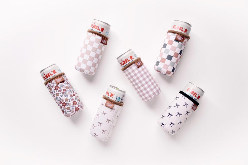 May include: Six beverage can coolers in various patterns, including checkered, floral, and bow designs. Each cooler has a brown band at the top and is filled with a can. The coolers are arranged on a white surface.