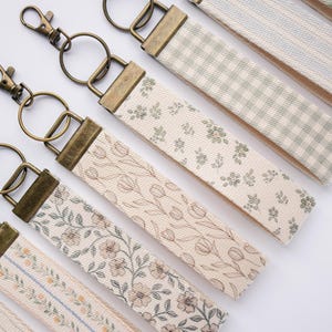 May include: A collection of fabric keychains with various floral and patterned designs. Each keychain features a brass-colored clasp and ring. The fabric straps display patterns like tulips, floral sprigs, and gingham checks in neutral tones.