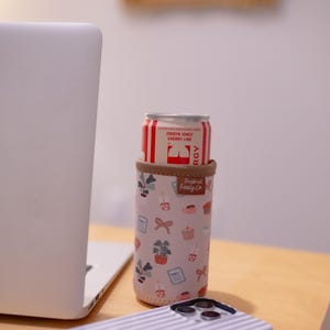 May include: A can of Jordyn Jones Cherry Lime soda in a pink and beige insulated beverage holder with a botanical pattern. The holder has a brown trim and the text "Frederick Family Co."
