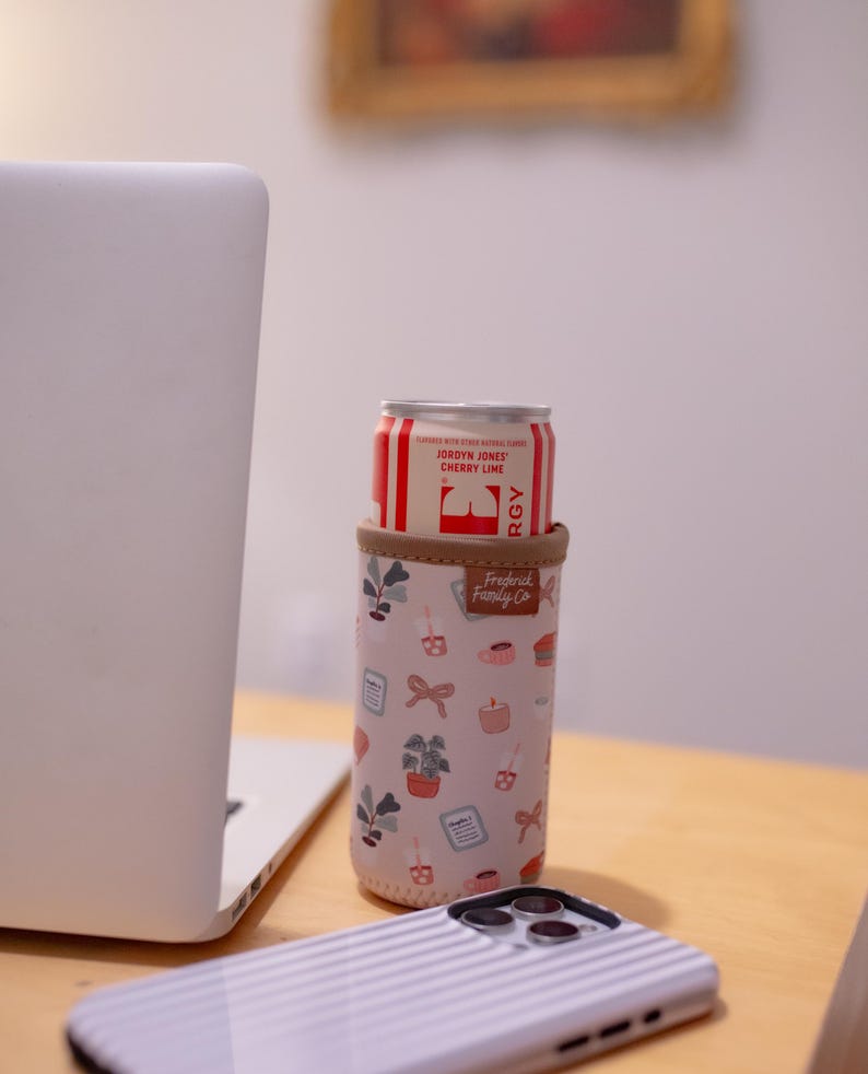 May include: A can of Jordyn Jones Cherry Lime soda in a pink and beige insulated beverage holder with a botanical pattern. The holder has a brown trim and the text "Frederick Family Co."