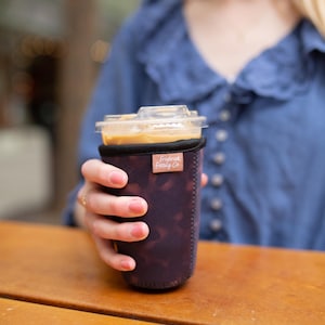 May include: A dark purple neoprene iced coffee cup sleeve with a subtle mottled pattern and a small rectangular tan fabric tag featuring the text Frederick Family Co. in white script, wrapped around a clear plastic cup filled with iced coffee.