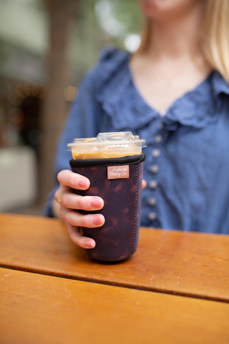 May include: A dark purple neoprene iced coffee cup sleeve with a subtle mottled pattern and a small rectangular tan fabric tag featuring the text Frederick Family Co. in white script, wrapped around a clear plastic cup filled with iced coffee.