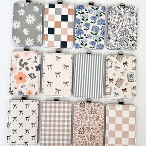 Puede incluir: Una colección de porta-credenciales rectangulares con varios diseños. Los diseños incluyen margaritas, patrones de cuadros, estampados florales, lazos, rayas y un estampado temático del desierto. Cada porta-credencial tiene un lazo metálico plateado en la parte superior.