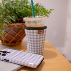 May include: A gray and white gingham patterned cup sleeve with a small brown rectangular tag labeled "Fredrick Family Co." wrapped around a clear plastic cup containing a light brown beverage and a teal straw, next to a matching gingham phone case.