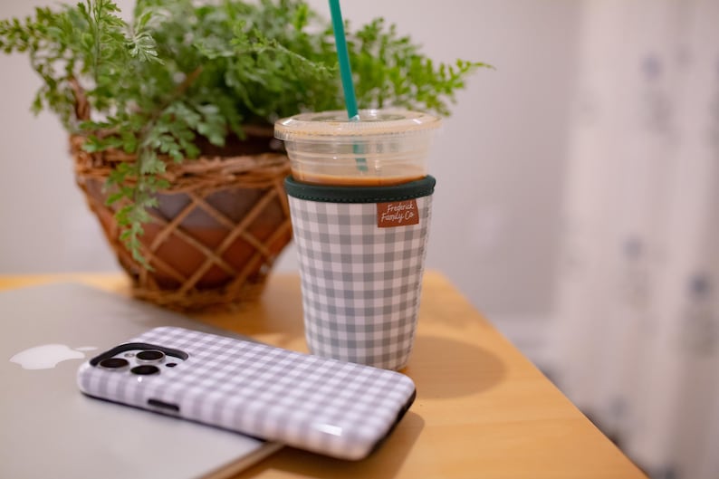 May include: A gray and white gingham patterned cup sleeve with a small brown rectangular tag labeled "Fredrick Family Co." wrapped around a clear plastic cup containing a light brown beverage and a teal straw, next to a matching gingham phone case.