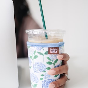 May include: A clear plastic iced coffee cup with a dark green straw, encased in a fabric sleeve featuring a white background with a blue and green hydrangea floral pattern and a brown rectangular tag labeled Frederick FAMILY.