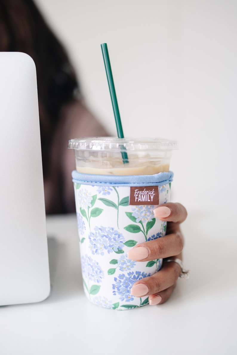 May include: A clear plastic iced coffee cup with a dark green straw, encased in a fabric sleeve featuring a white background with a blue and green hydrangea floral pattern and a brown rectangular tag labeled Frederick FAMILY.