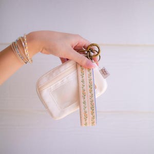 May include: A cream-colored zippered pouch with a clear ID window and a fabric wrist strap featuring a floral pattern of small orange flowers and green leaves. The pouch includes a bronze-toned metal keyring and a small white tag labeled Threaded Fairy Co.
