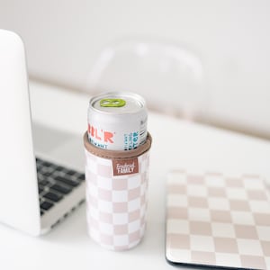 May include: A silver can of sparkling water sits in a pink and white checkered can cooler with a brown band. The cooler has the text "Frederick Family" on it. A matching checkered coaster is next to the cooler.