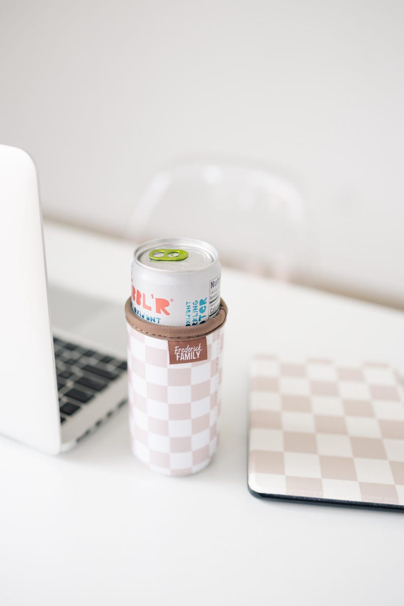 May include: A silver can of sparkling water sits in a pink and white checkered can cooler with a brown band. The cooler has the text "Frederick Family" on it. A matching checkered coaster is next to the cooler.