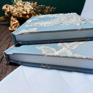 May include: Two blue journals with white floral embroidery on the covers. The journals are stacked on top of each other.