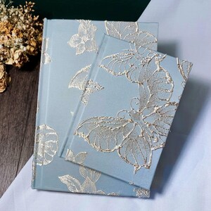 May include: Two blue journals with a silver butterfly and floral pattern. The smaller journal is on top of the larger journal.