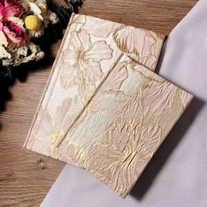 Puede incluir: Dos diarios con estampado floral rosa y dorado. Los diarios están hechos de una tela suave y texturizada.