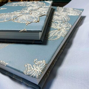 May include: Two blue journals with white and gold floral embroidery. The journals are stacked on top of each other.