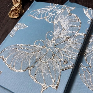May include: A light blue notebook with a silver butterfly design. The butterfly is embroidered on the cover of the notebook.