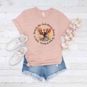 May include: A pink t-shirt with a colorful phoenix design and the text "The Spirit Of Heaviness - Isaiah 61:3 - Beauty for Ashes. Joy for Mourning. Praise for the Spirit Of Heaviness."