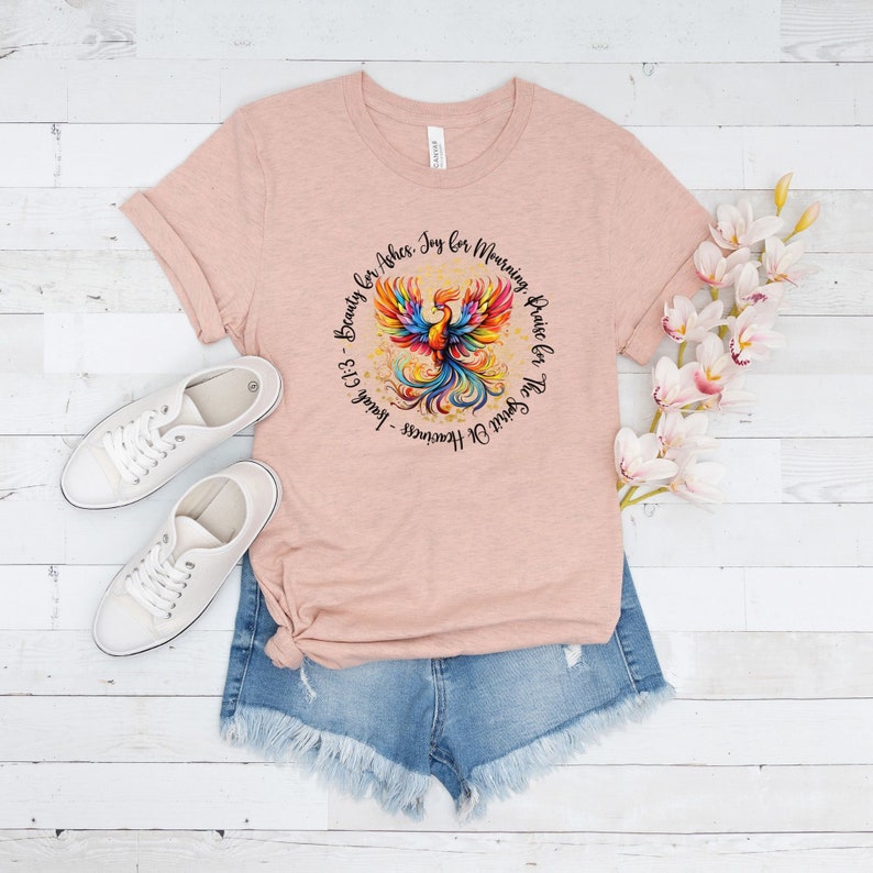 May include: A pink t-shirt with a colorful phoenix design and the text "The Spirit Of Heaviness - Isaiah 61:3 - Beauty for Ashes. Joy for Mourning. Praise for the Spirit Of Heaviness."