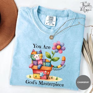 May include: A light blue crew neck t-shirt featuring a graphic of a patchwork cat sitting in a flower pot next to a colourful flower. The text reads You Are God's Masterpiece and Ephesians 2:10. The shirt has a Comfort Colors brand label.