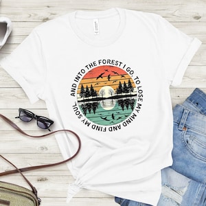May include: White t-shirt with a circular graphic featuring a sunset over a lake with trees and birds. The text around the graphic reads, "And into the forest I go, to lose my mind and find my soul."